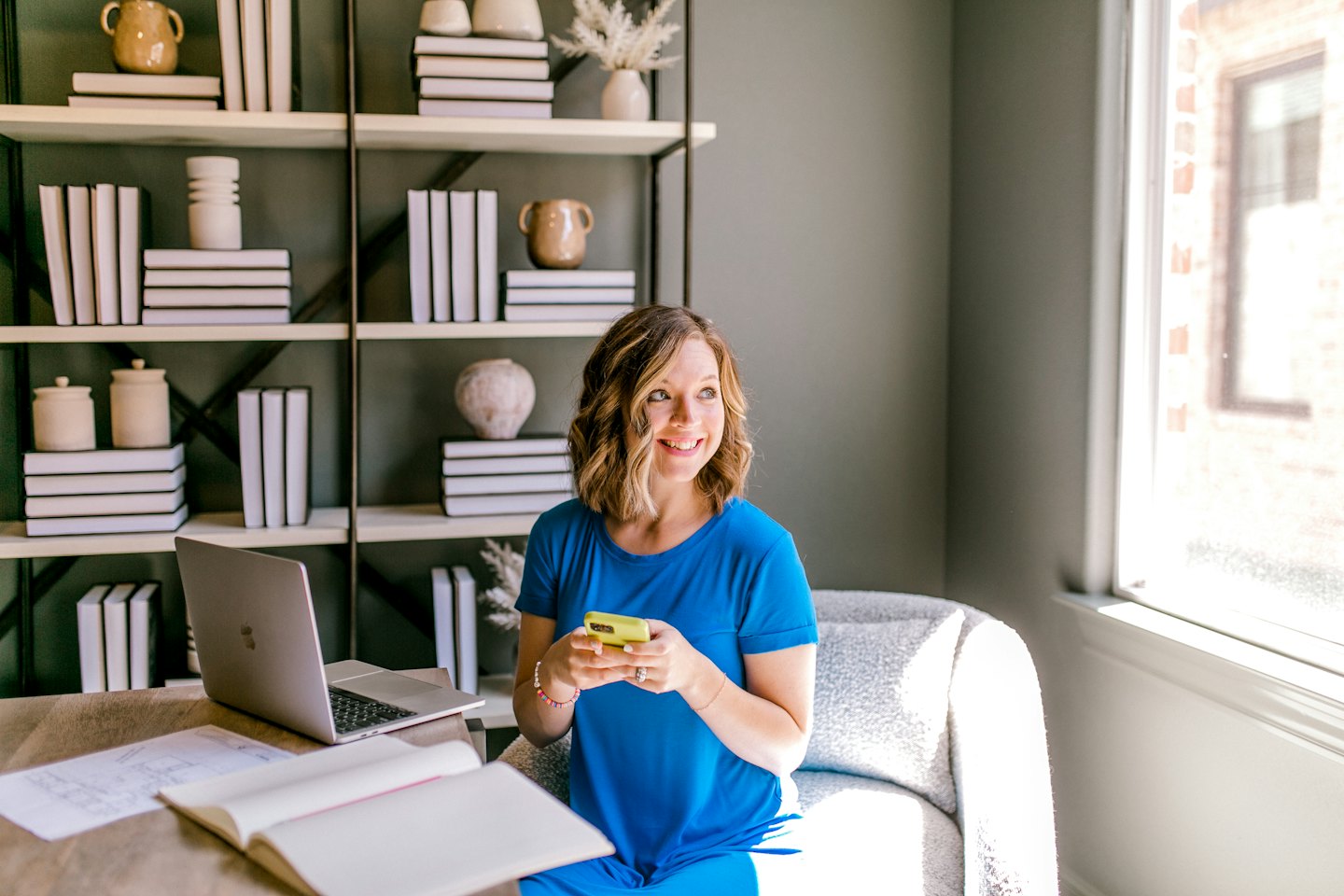 Frau im blauen Kleid benutzt Smartphone am Schreibtisch mit Laptop und Bücherregal.